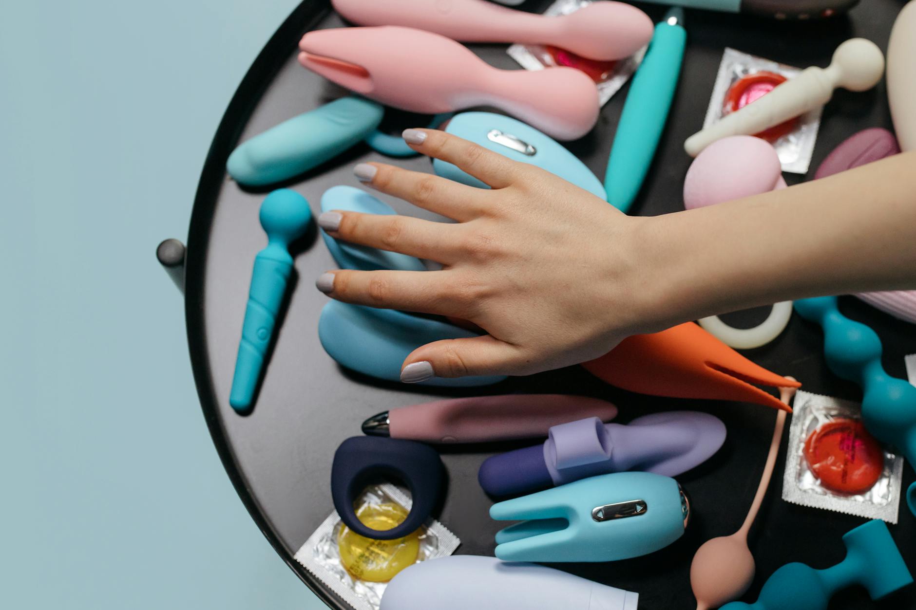 A hand reaching over a variety of colorful vibrators on a table, showing multiple intimate wellness options