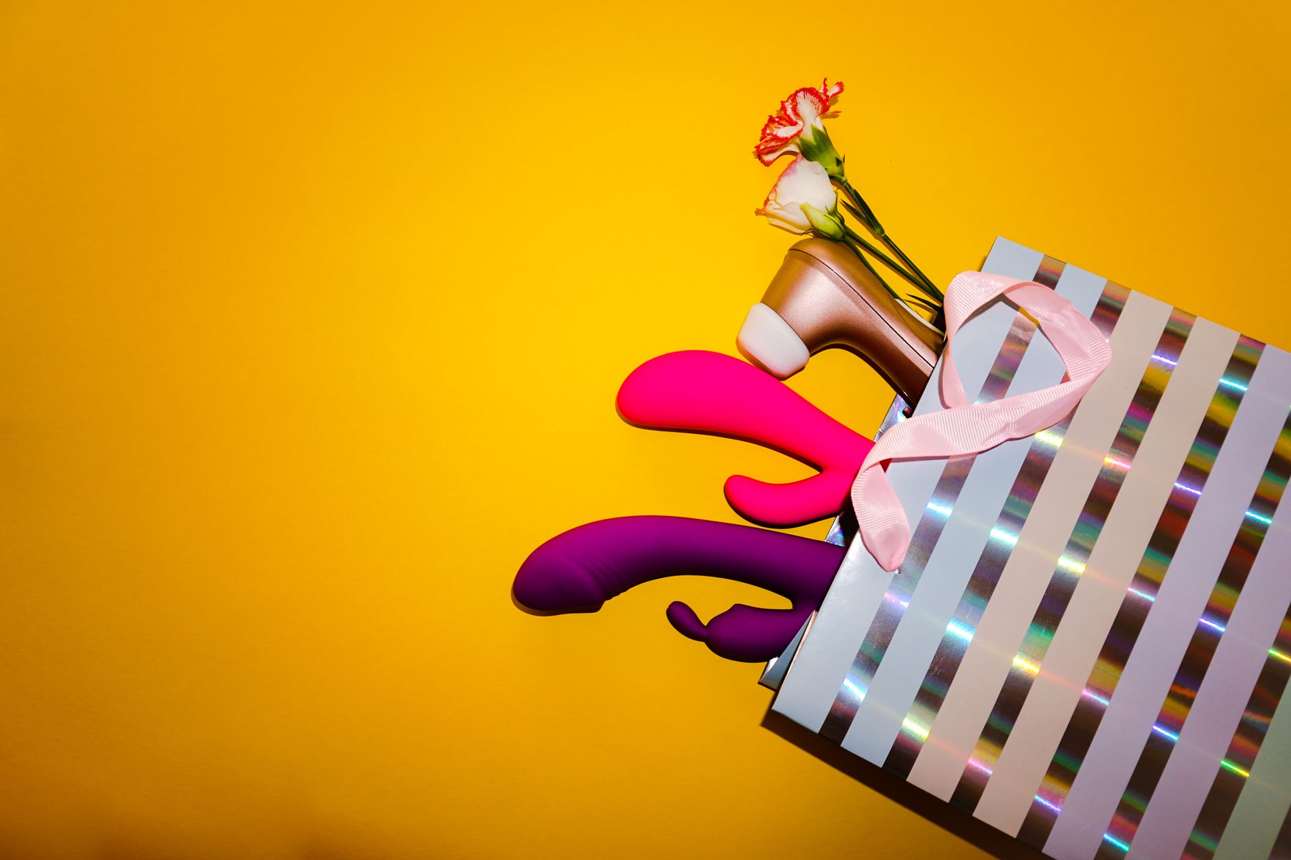 Colorful vibrators with flowers in a holographic gift bag against yellow background